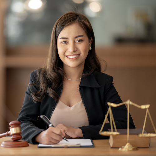 An attorney sits at a desk, pen poised for writing in her notebook, with a judge’s gavel and a set of brass scales on the desk near her.