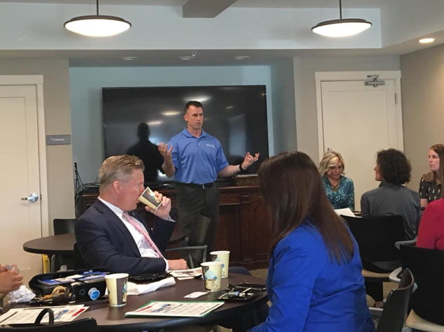 A man in a blue shirt stands before a blank black screen presenting St. Joseph printing services to a group in a conference room.