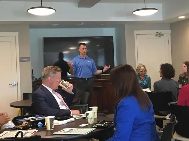 A man in a blue shirt stands before a blank black screen presenting St. Joseph printing services to a group in a conference room.